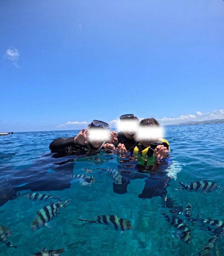 沖縄の青い海でシュノーケリング中に、周囲を泳ぐ熱帯魚と一緒にシーサーポーズで記念撮影をする家族。