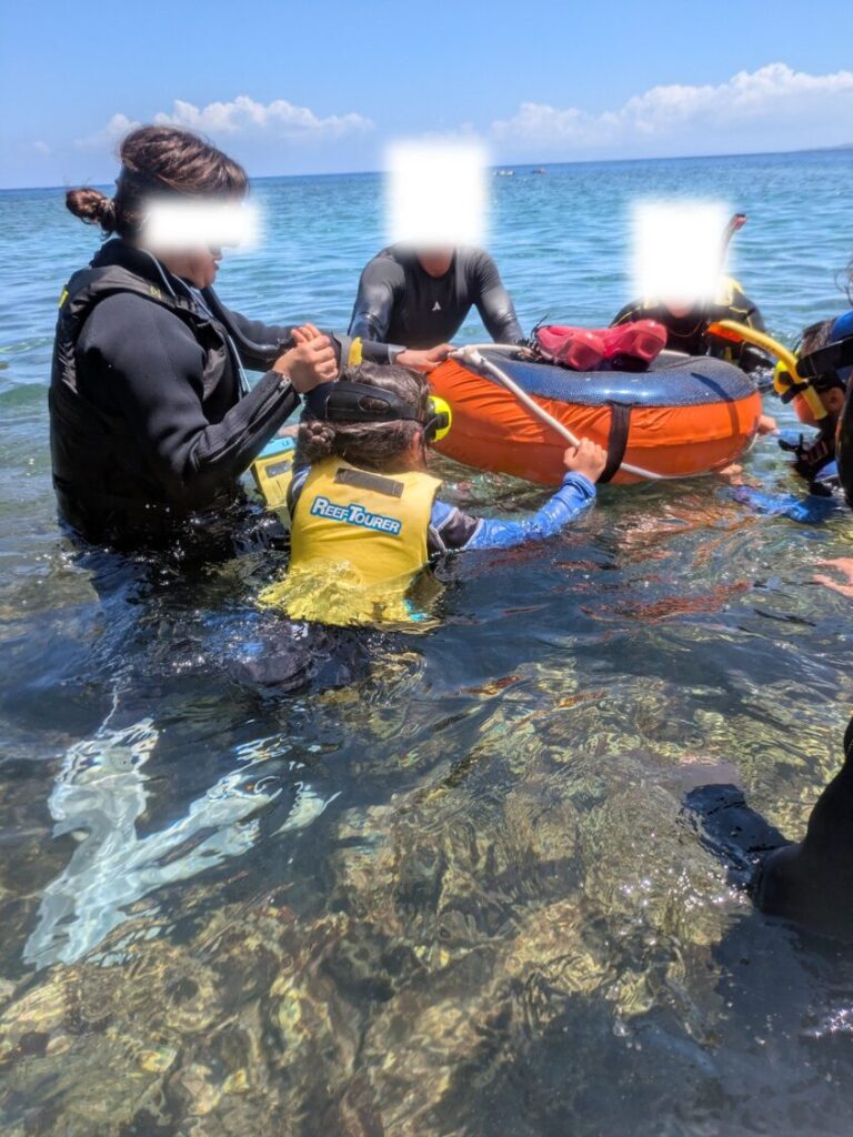 浅瀬の海でオレンジ色の浮き輪につかまり、ガイドにシュノーケルマスクの調節をしてもらいながら安心して海に入る準備をする子供と家族。