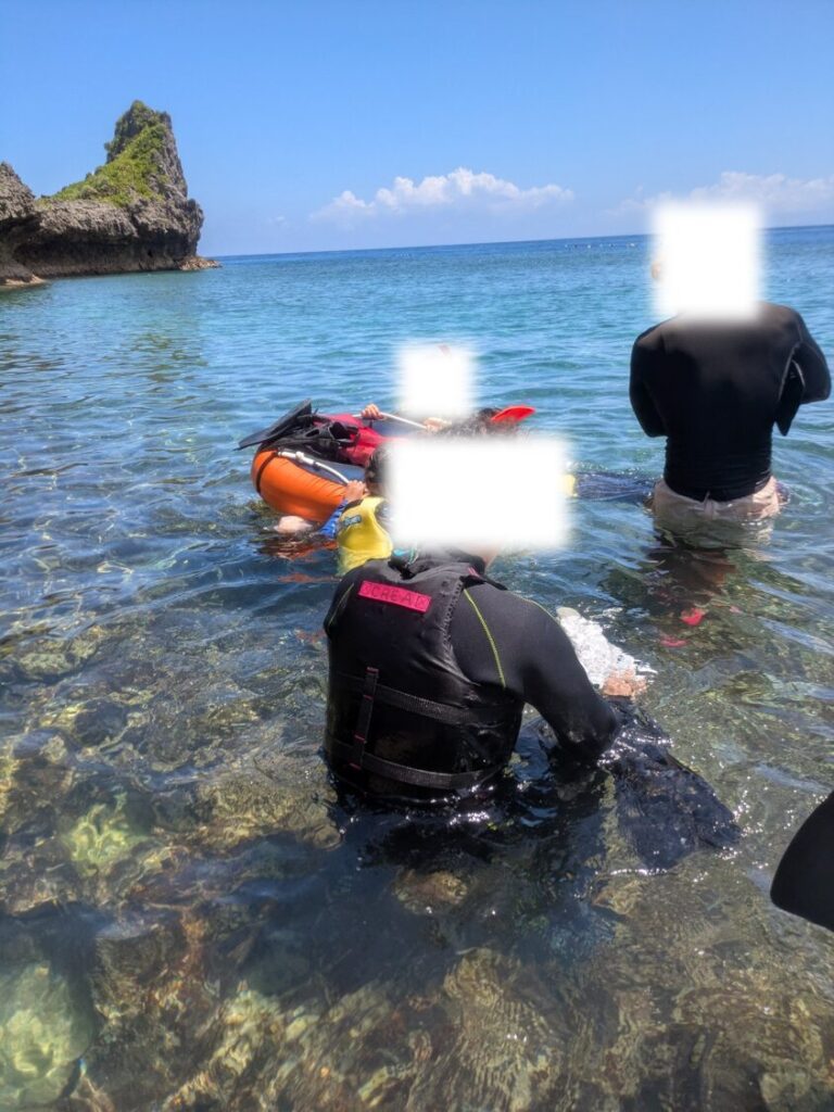 青の洞窟前の浅瀬で海に入り、ガイドにフィンの装着を手伝ってもらいながら浮き輪につかまってシュノーケリングの準備をする家族。