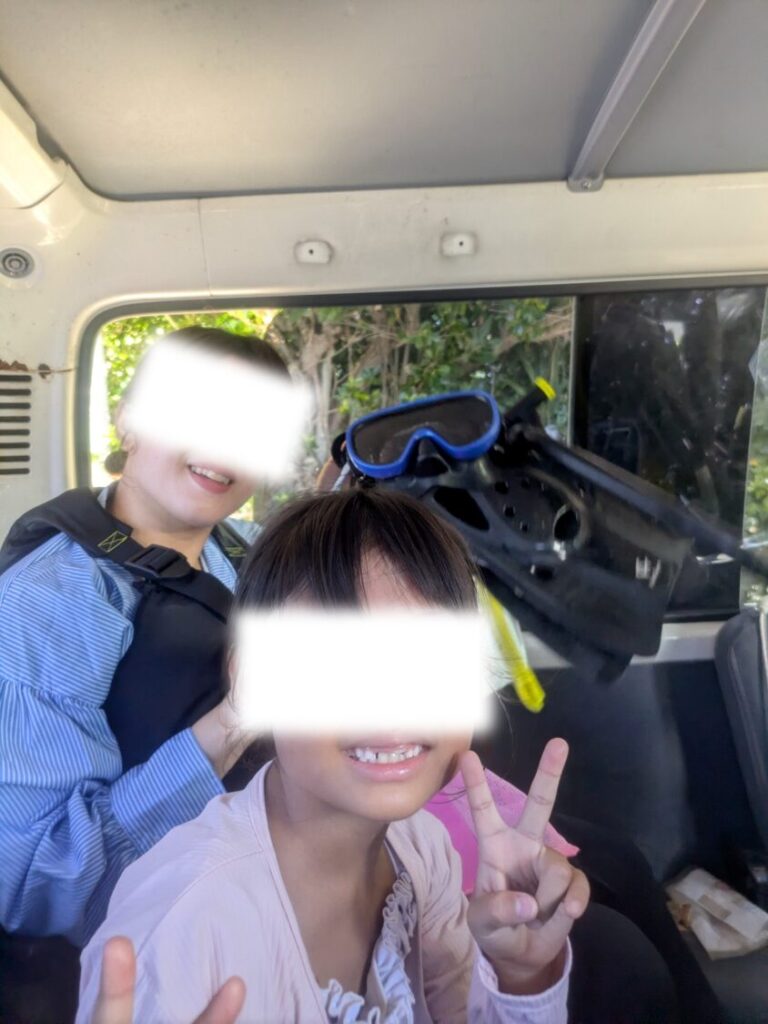 青の洞窟へ向かう送迎車の車内で、シュノーケルメガネとフィンを手に笑顔でピースサインをする親子。