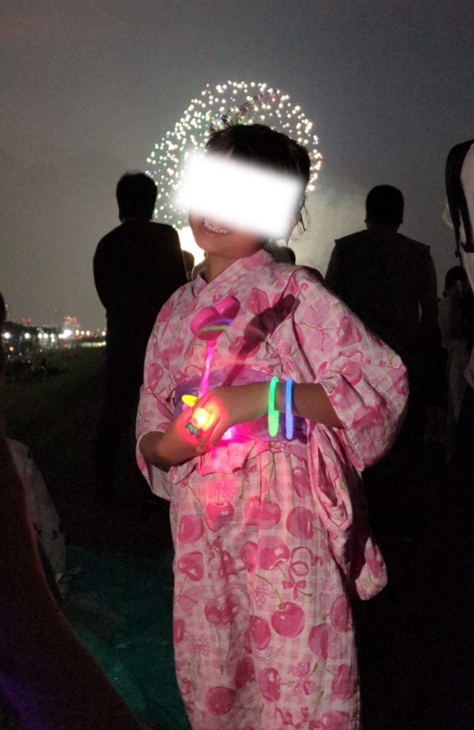 打ち上がる花火を背景に光るグッズを手にして微笑む浴衣姿の子供