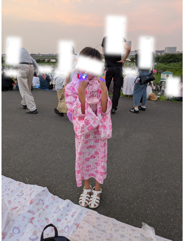 江戸川河川敷の土手手前側にレジャーシートを敷き、ピースサインをする浴衣姿の子供