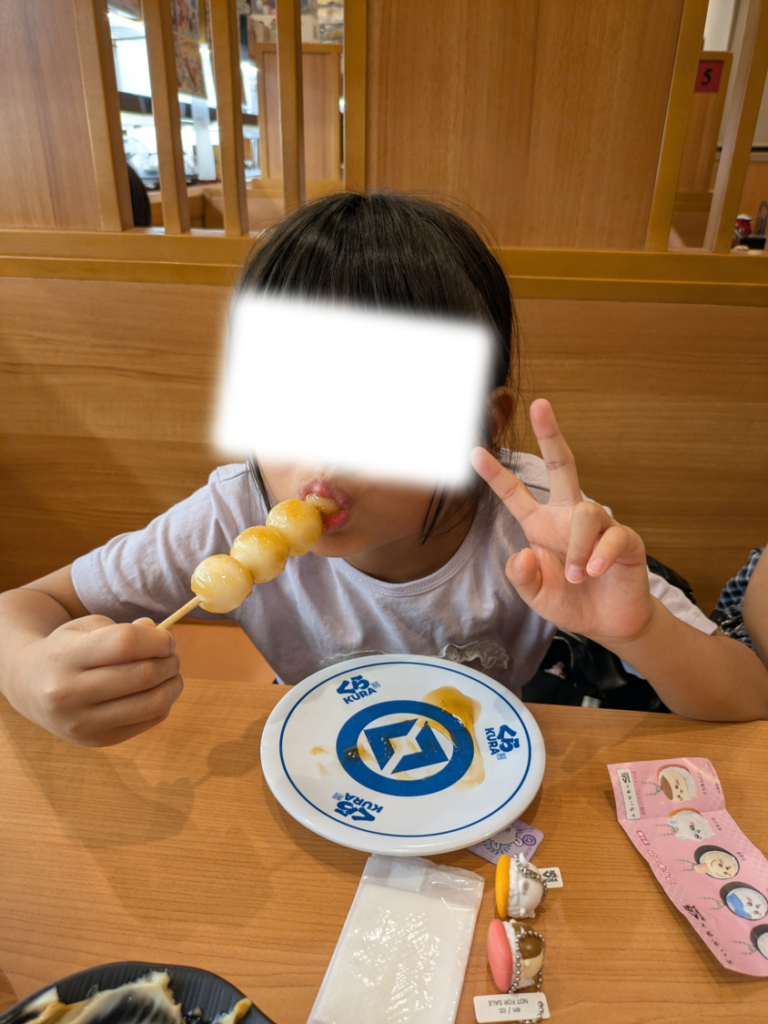 くら寿司で平皿に乗ったみたらし団子を食べる子どもと、テーブルの上に置かれたビッくらポンの景品