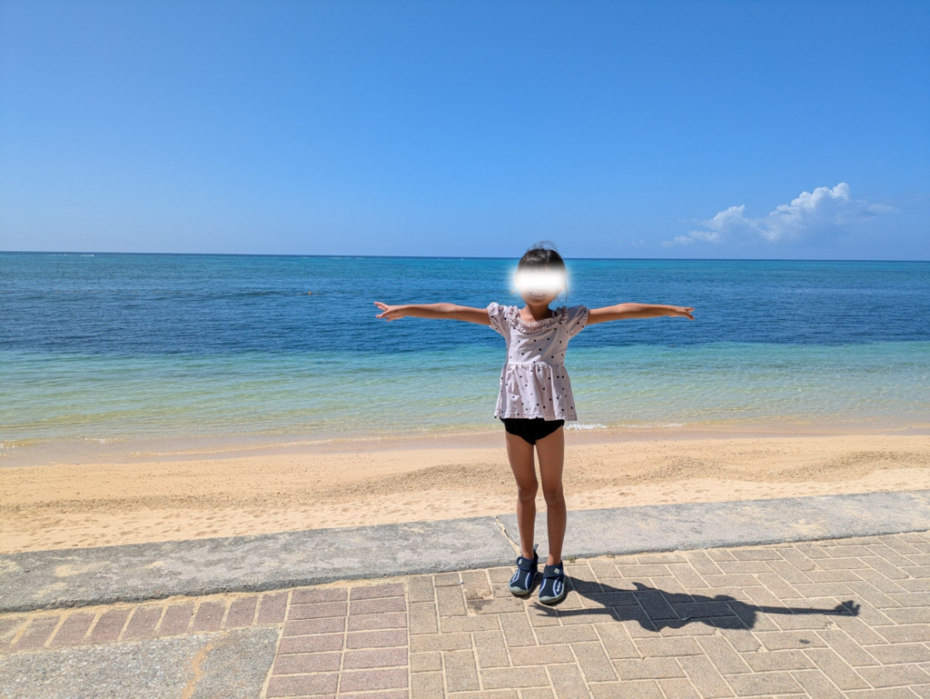沖縄の美しいビーチを背景に、マリンシューズを履いて両腕を大きく広げる笑顔の娘。
