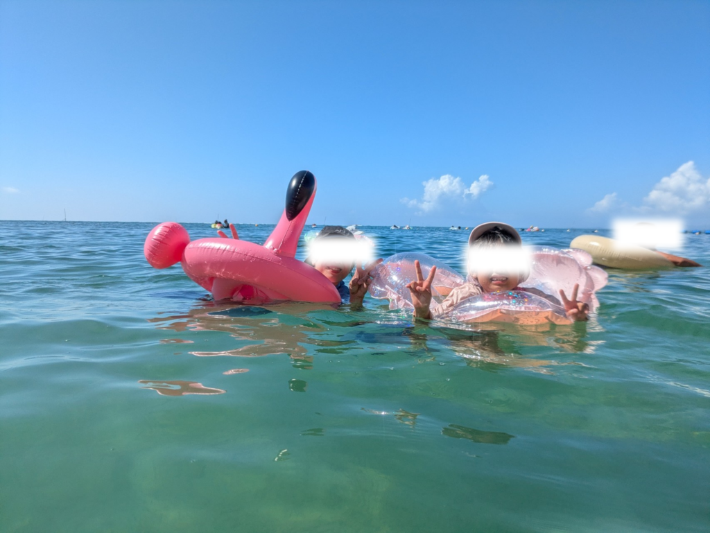 沖縄の澄んだ海で浮き輪に乗りピースをする子供。表情は楽しそうだが「しょっぱい！」と海水の塩分を気にしている様子。