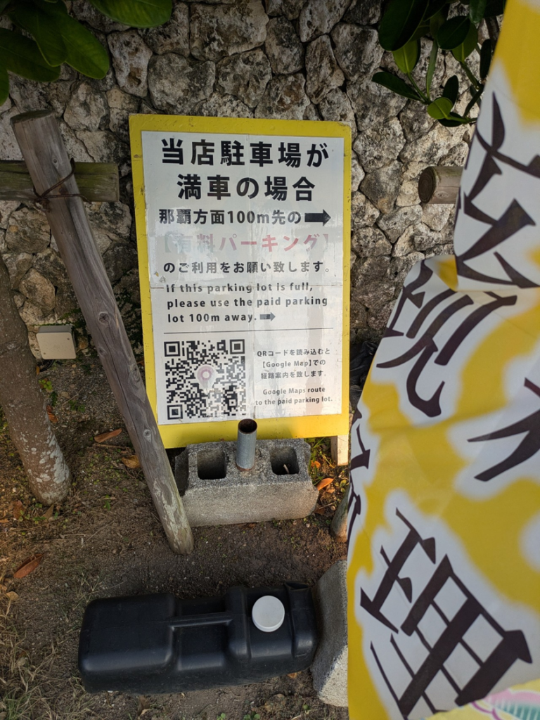 沖縄の台所 ぱいかじ恩納本店の駐車場満車案内看板。那覇方面へ100m先の有料パーキング利用を促す説明と、Googleマップ用QRコードが記載されている。