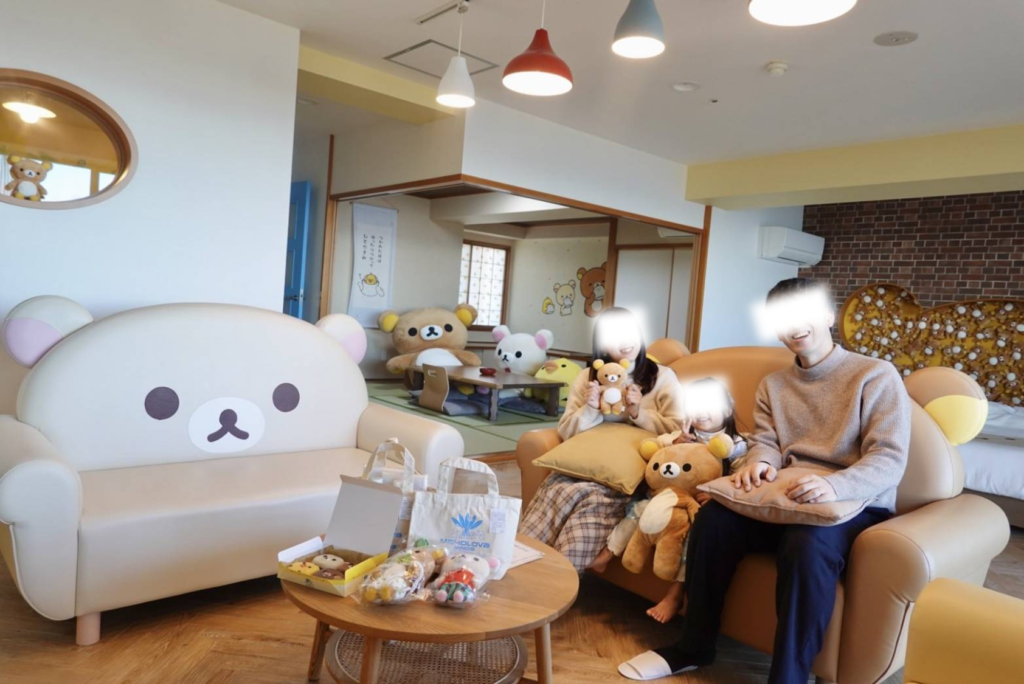 マホロバ・マインズ三浦のリラックマルーム。リラックマやコリラックマの大きなソファがあるリビングで、ぬいぐるみを手に笑顔でくつろぐ家族の記念写真