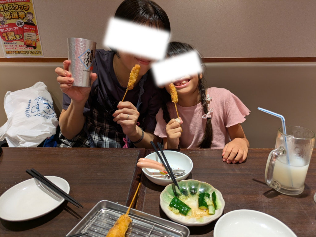 「新世界串カツ いっとく 三番街店」の店内で、串カツを手に持って嬉しそうにポーズをとる様子